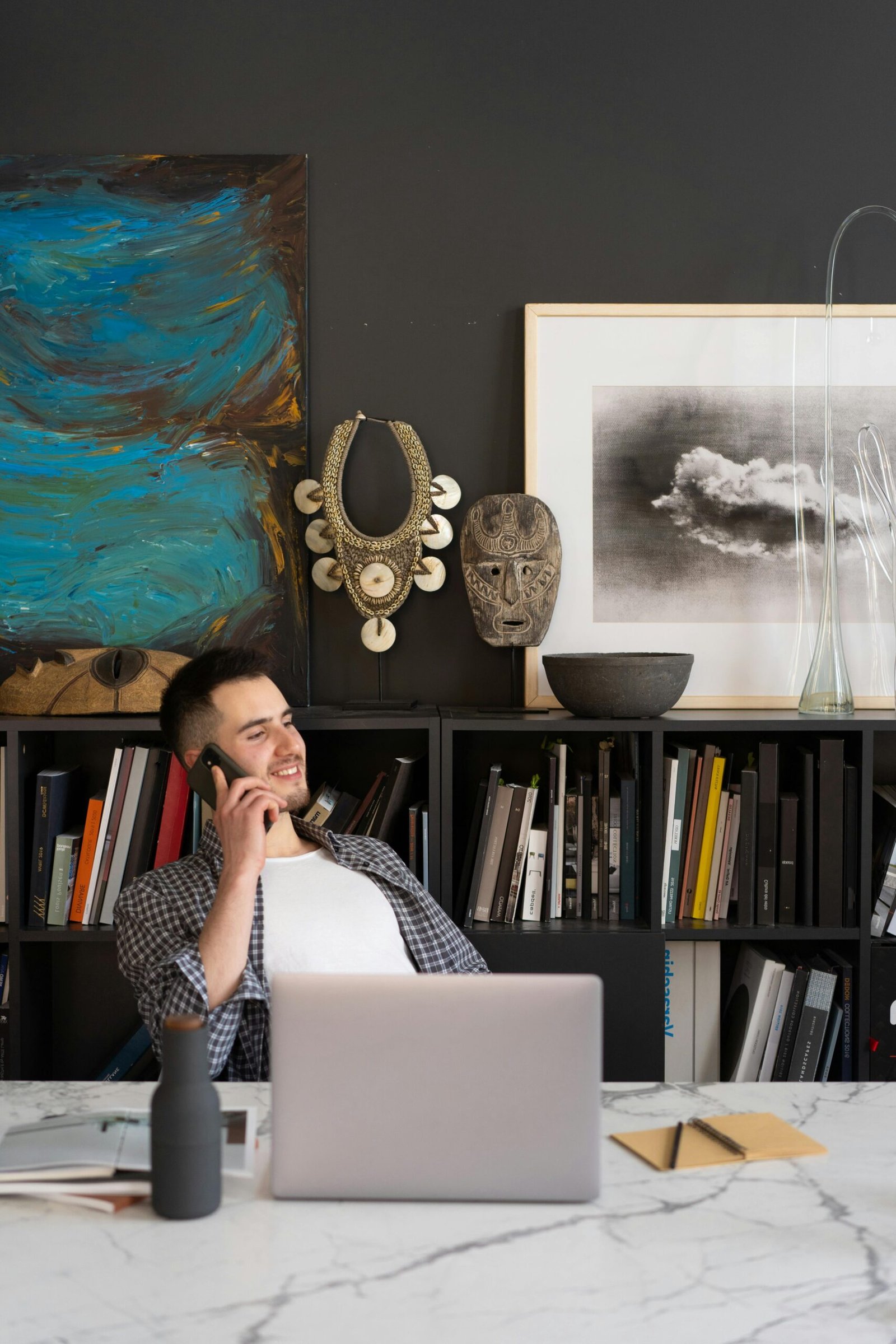 Man talking on phone in modern home office, surrounded by artwork and books.