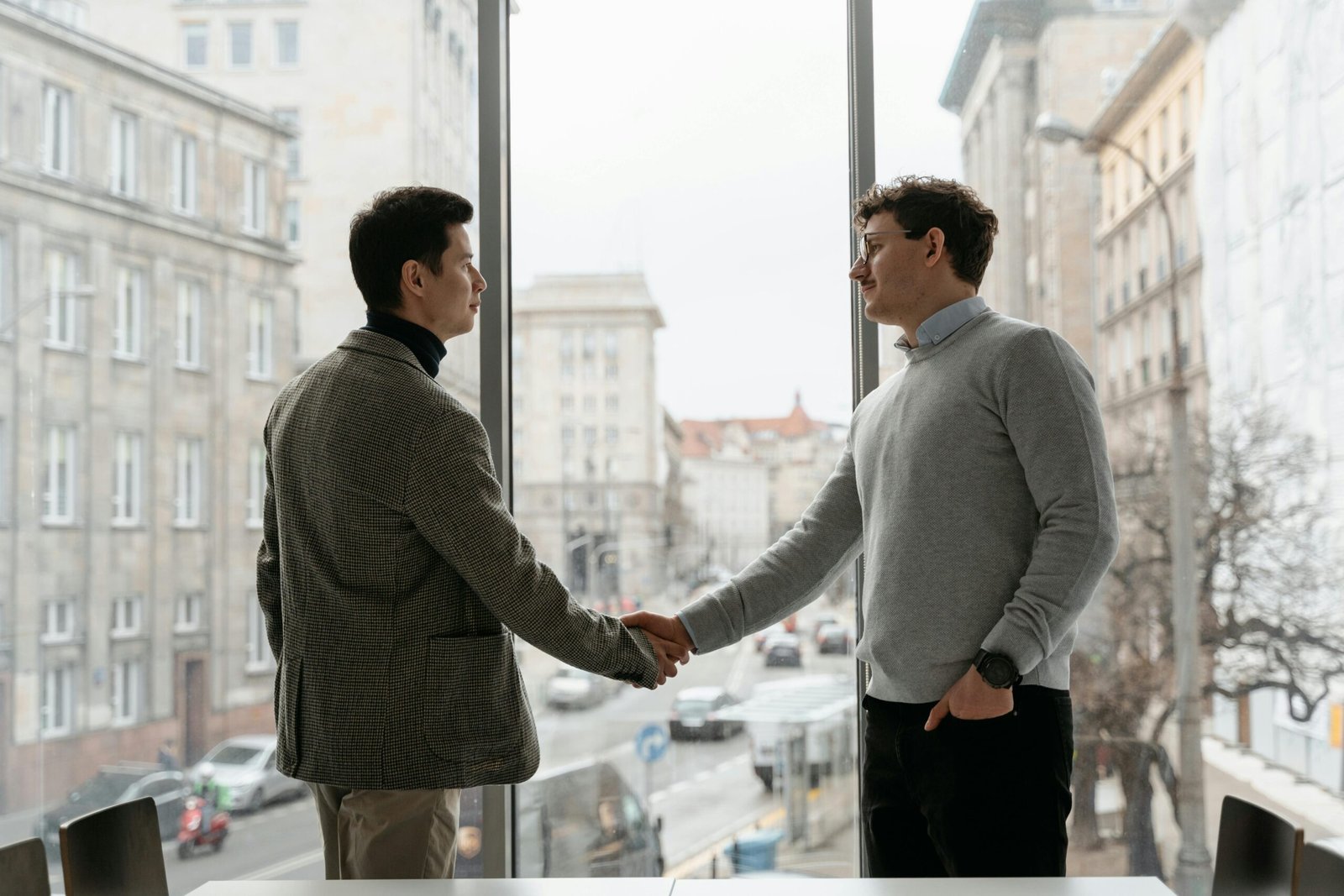 Two businessmen shaking hands in an urban office with city view through large windows.
